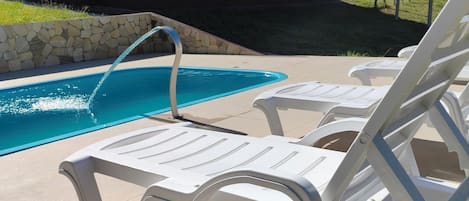 A pool area with white lounge chairs, a stone wall, and a house in the background.