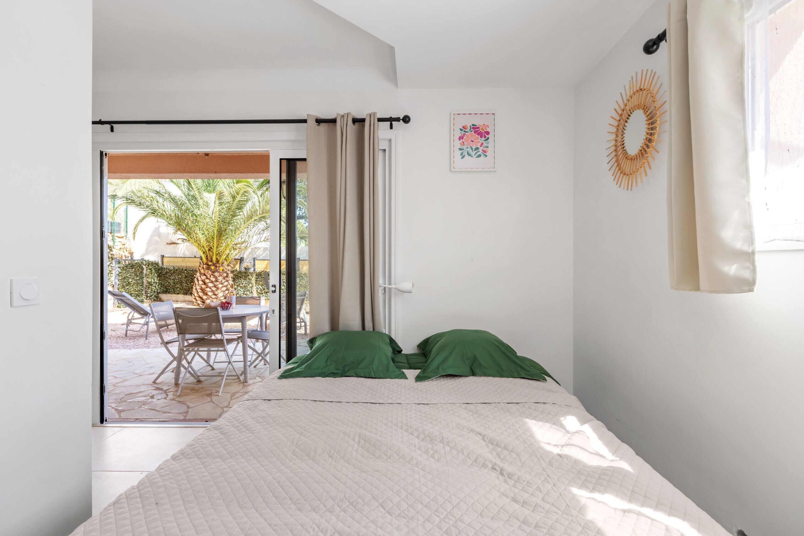 Un dormitorio con cama, almohadas verdes, ventana con cortinas, decoración en la pared y un patio exterior con mesa y sillas.