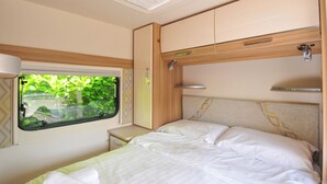 A compact RV bedroom with a bed, a window with a view of greenery, and a ceiling fan. - Rod and Rest (Chulmleigh)