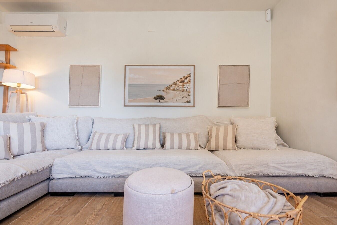 Een woonkamer met een hoekbank, een rond voetenbankje en een rieten fauteuil. Drie ingelijste kunstwerken hangen aan de muur en links staat een lamp.
