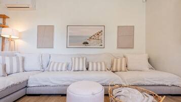 Een woonkamer met een hoekbank, een rond voetenbankje en een rieten fauteuil. Drie ingelijste kunstwerken hangen aan de muur en links staat een lamp.