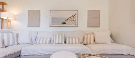 Un salón con un sofá seccional, un otoman redondo y una silla de mimbre. Hay tres obras de arte enmarcadas en la pared y una lámpara a la izquierda.
