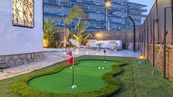 Een minigolfbaan met een rode vlag, witte lounge fauteuils en een zwembad op de achtergrond.