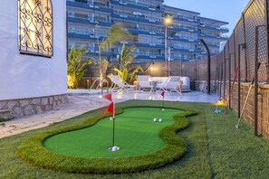 Minigolfrata, jossa on punainen lippu, valkoiset aurinkotuolit ja taustalla uima-allas.