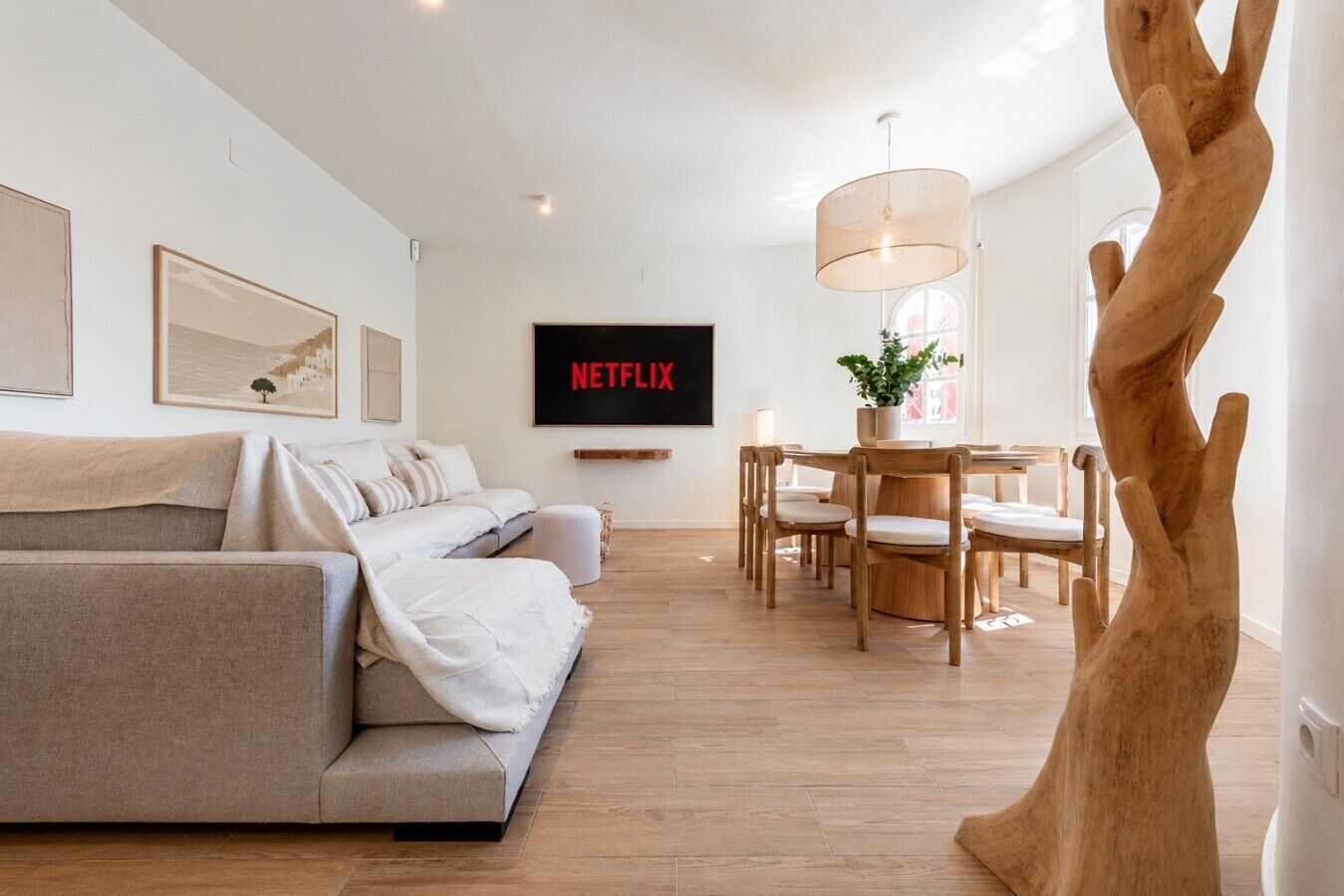 Un salon moderne comprenant un canapé, une table à manger avec des chaises, une sculpture en bois représentant un arbre et un téléviseur fixé au mur qui affiche « NETFLIX ».