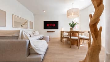 Een moderne woonkamer met een bank, een eettafel met stoelen, een houten boomsculptuur en een aan de muur bevestigde televisie waarop 'NETFLIX' wordt weergegeven.