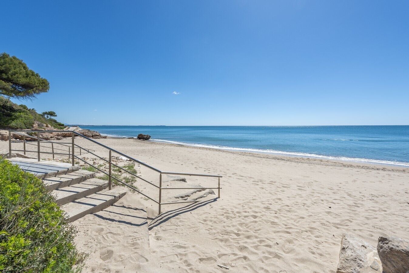 Een strand met trappen die naar het strand leiden, een heldere blauwe lucht en een rustige zee.