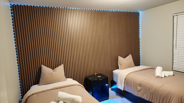 Een slaapkamer met twee bedden, een nachtkastje en een wand met verticale lamellen.