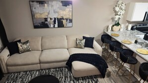 Een woonkamer met een bank, een zwart-wit tapijt met patroon, een keuken met een aanrecht, barkrukken en een decoratieve plant.
