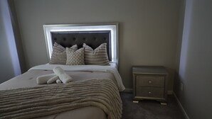 Een net opgemaakt bed met een gestoffeerd hoofdeinde, een nachtkastje met een structuur en een vloerbedekking.