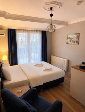 Habitación de hotel con cama, escritorio, silla, ventana con cortinas, radiador y un cuadro en la pared.