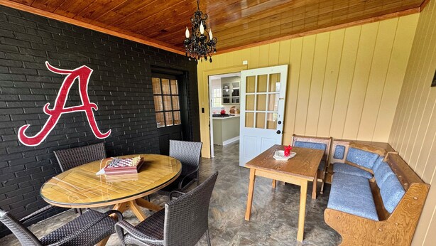 Ein Zimmer mit schwarzer Ziegelwand, Holzboden, einem runden Tisch mit Schachspiel, Korbstühlen, einer Holzbank mit blauen Kissen und einer zur Küche führenden Tür.