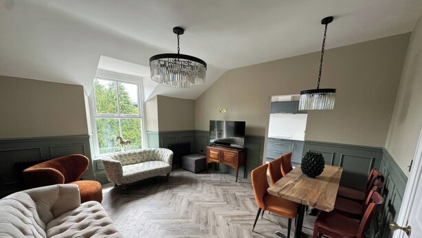 Ein Zimmer mit Fischgrätparkett, einem gesteppten Sofa, einem Esstisch mit Stühlen und einem Fernseher.