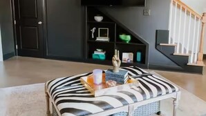 Un salon avec un canapé noir, un téléviseur à écran plat fixé au mur, un escalier et une bibliothèque contenant divers objets.