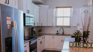 Une cuisine moderne avec des meubles blancs, un réfrigérateur en acier inoxydable, un four encastré et une fenêtre équipée de stores.