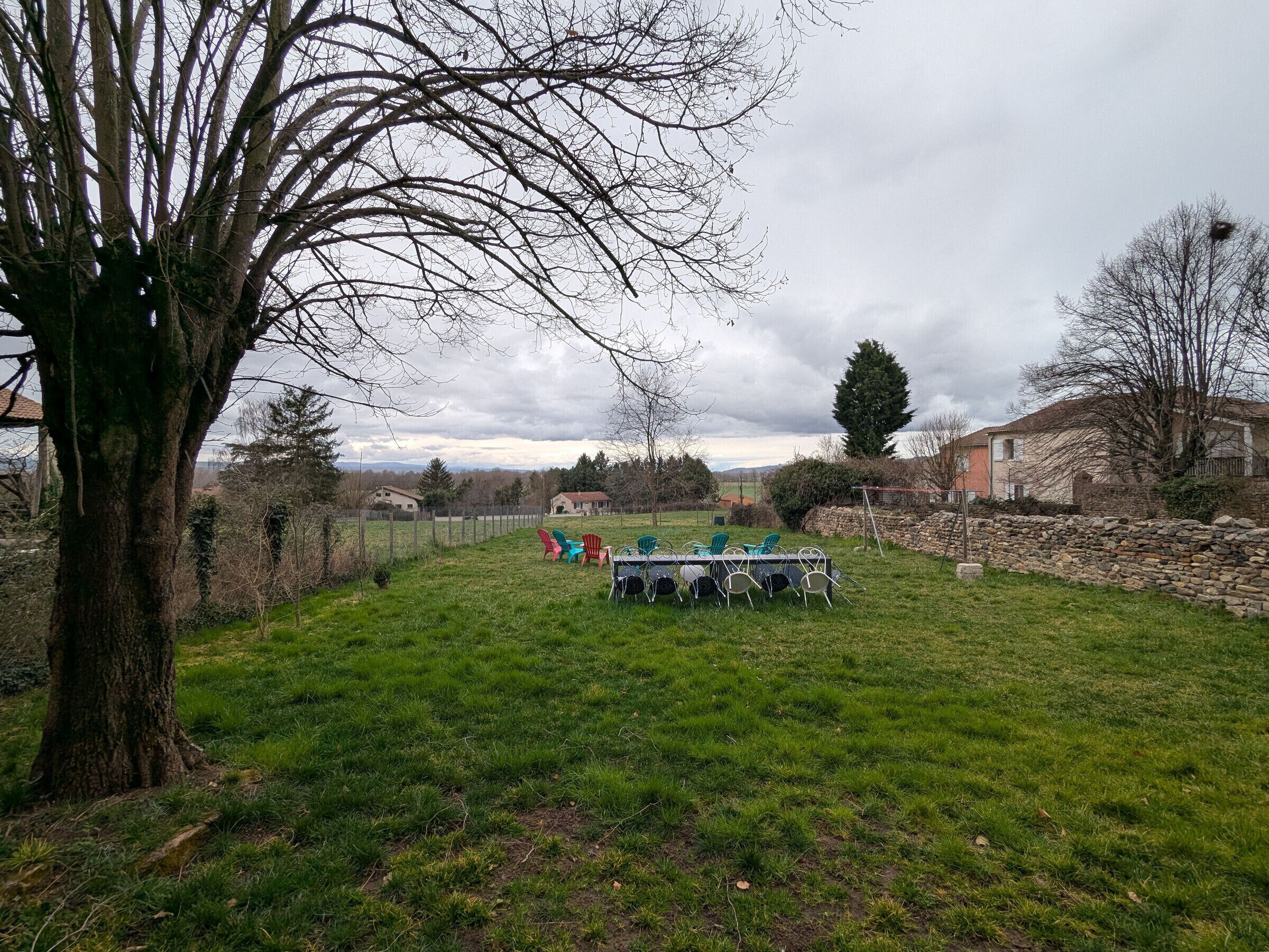 A grassy outdoor area with a table and chairs, surrounded by trees and a stone wall.
