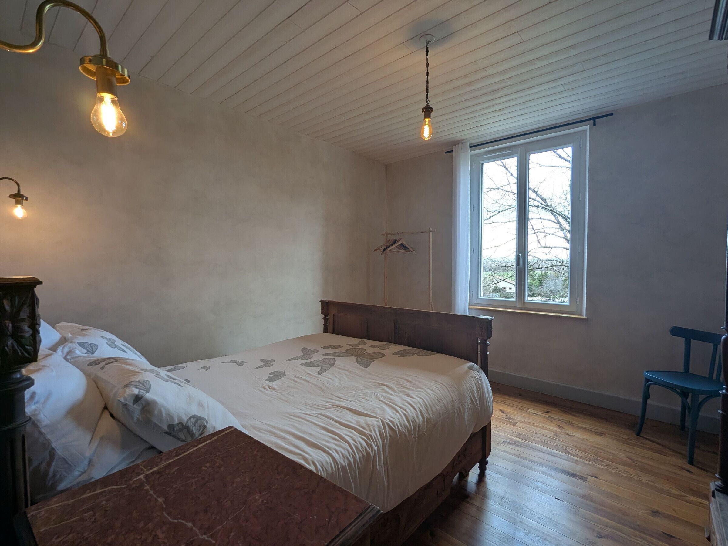Un dormitorio con una cama de madera, un banco de madera, una silla, un ventanal y dos lámparas colgantes.