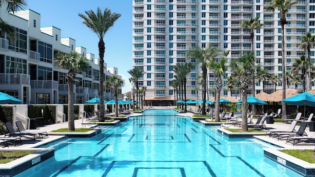 Ein Pool mit Liegestühlen und Sonnenschirmen, umgeben von Palmen und einem Hochhaus.