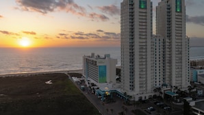 Ein Hochhaus-Hotel mit Blick auf den Sonnenuntergang über dem Meer, das mehrere Stockwerke und ein markantes Gebäude mit einer grün beleuchteten Spitze zeigt.