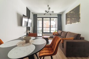 Un salon moderne avec un canapé marron, une table à manger avec des chaises, un téléviseur à écran plat fixé au mur et une grande fenêtre avec des rideaux.