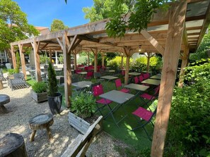 Un patio al aire libre con mesas y sillas, pérgola de madera y follaje.