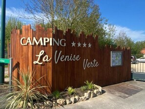 Ein hölzernes Schild mit der Aufschrift 'CAMPING La Venise Verde', umgeben von Pflanzen und blauem Himmel.