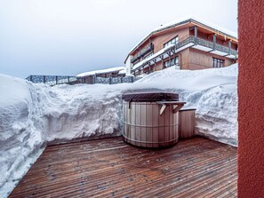 Eine Holzterrasse mit Sauna, verschneiter Umgebung und einem Gebäude mit Balkonen.