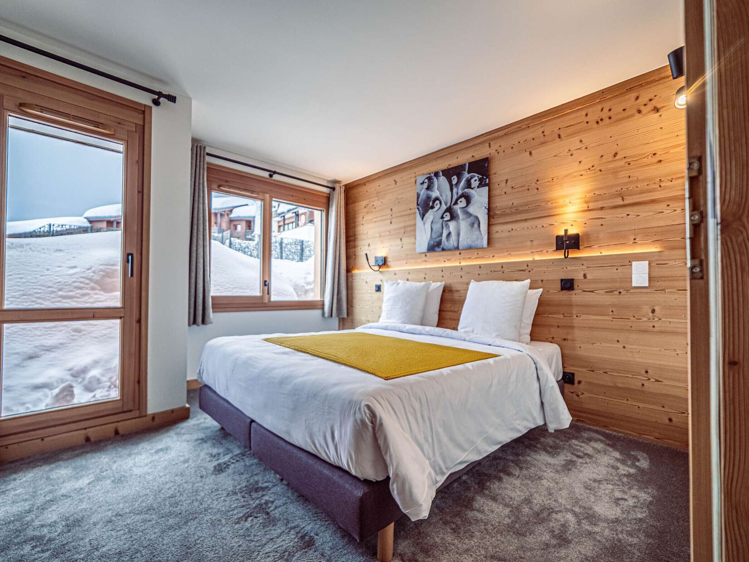 A bedroom with a bed, a window with a view of snow-covered buildings, wooden paneling on the walls, and a painting above the bed.