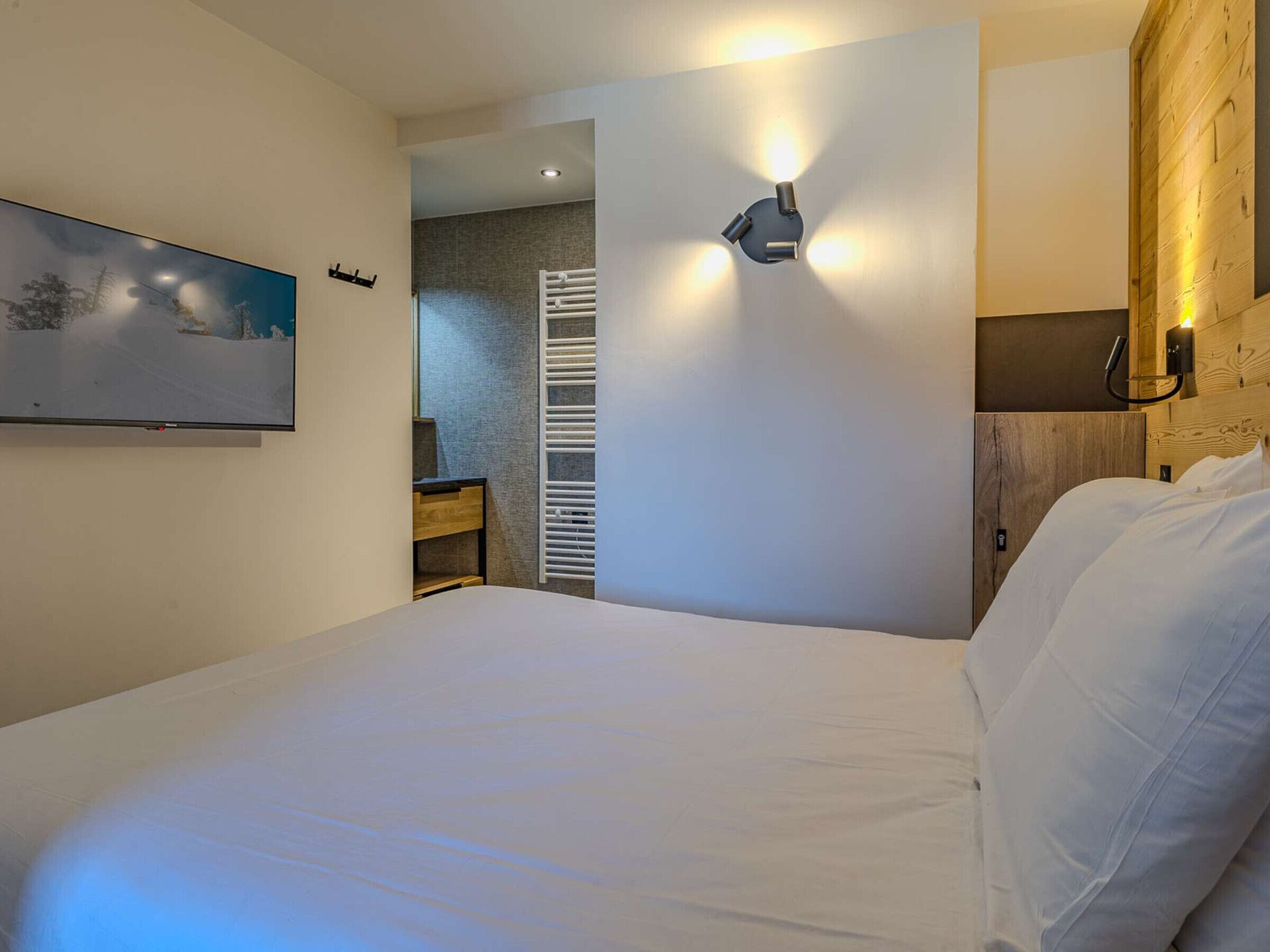 Une chambre à coucher comprenant un lit, un téléviseur fixé au mur, un radiateur intégré et une tête de lit en bois.