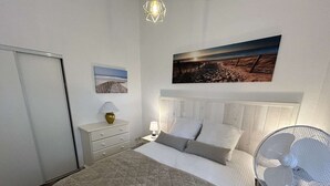 Et soveværelse med en seng, et natbord med en lampe, et billede hængt på væggen og en gulvventilator.