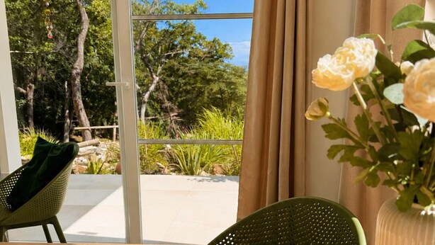 Quarto com uma mesa de madeira, jogos americanos trançados, um vaso com flores e vista para árvores e vegetação lá fora.