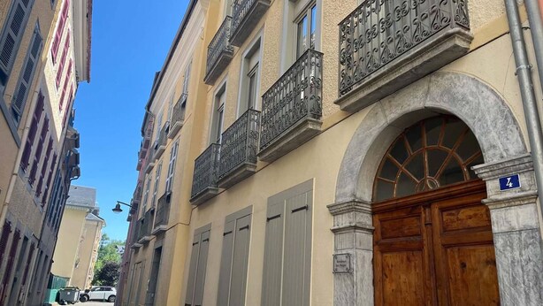 A narrow street with buildings featuring balconies, a wooden door, and a clear blue sky. - Bright, peaceful apartment - 8 guests (Cauterets)