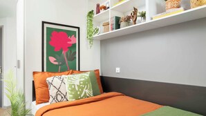 A bedroom with a bed, pillows, a framed picture, a shelf with books and plants, and a potted plant.