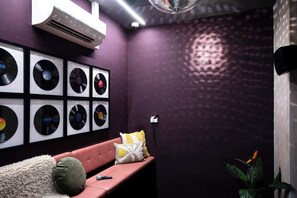A room with a purple wall, a framed vinyl record collection, a pink sofa, a microphone, and a plant.