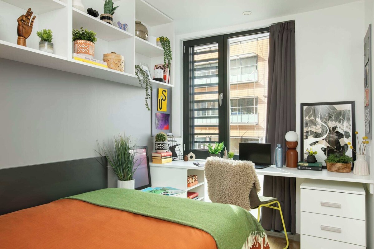 A modern bedroom with a bed, desk, chair, and shelving with plants and books.