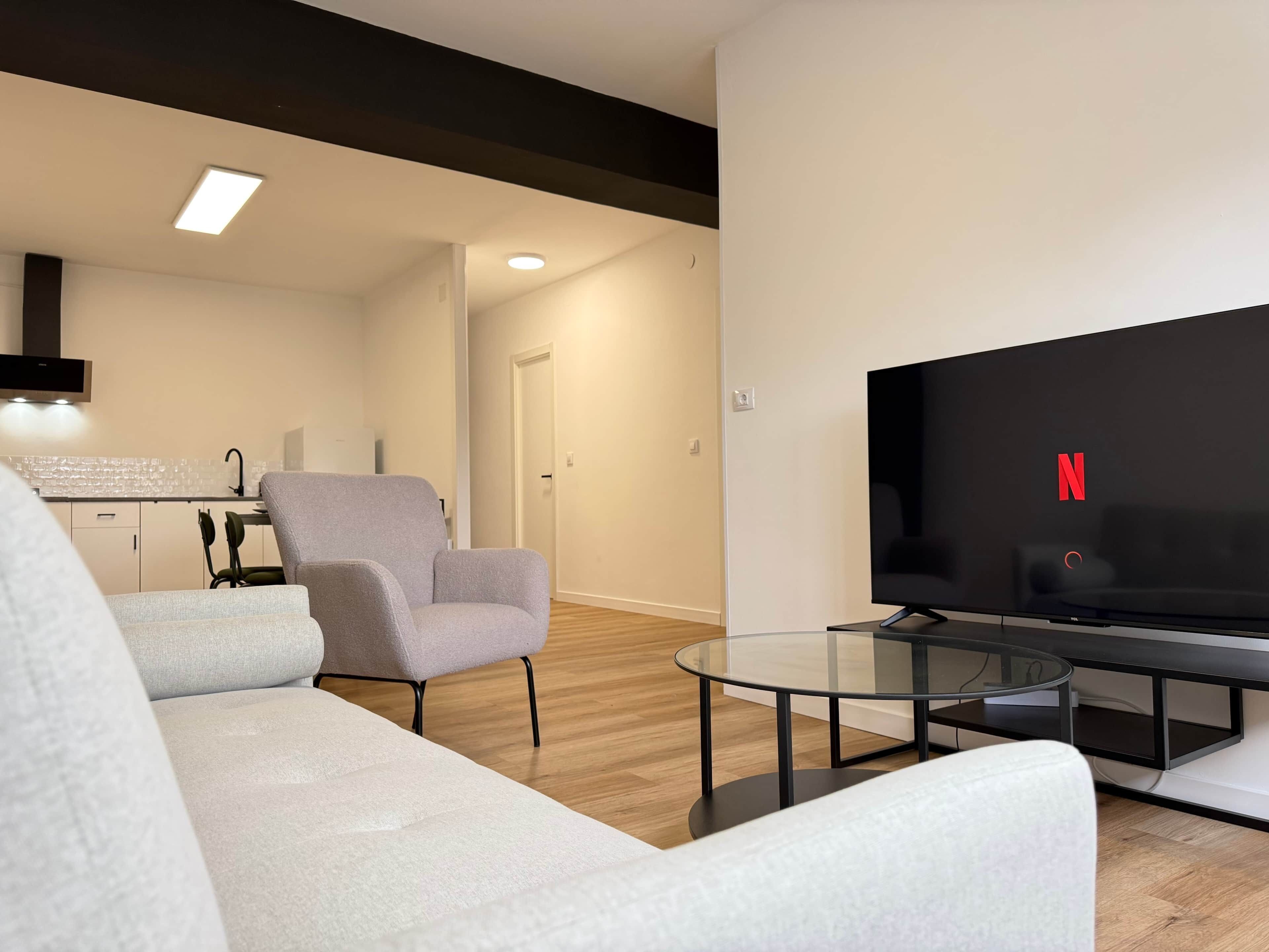 Una sala moderna con un sofá, un sillón, una mesa de centro de vidrio y un televisor de pantalla plana mostrando el logotipo de Netflix.