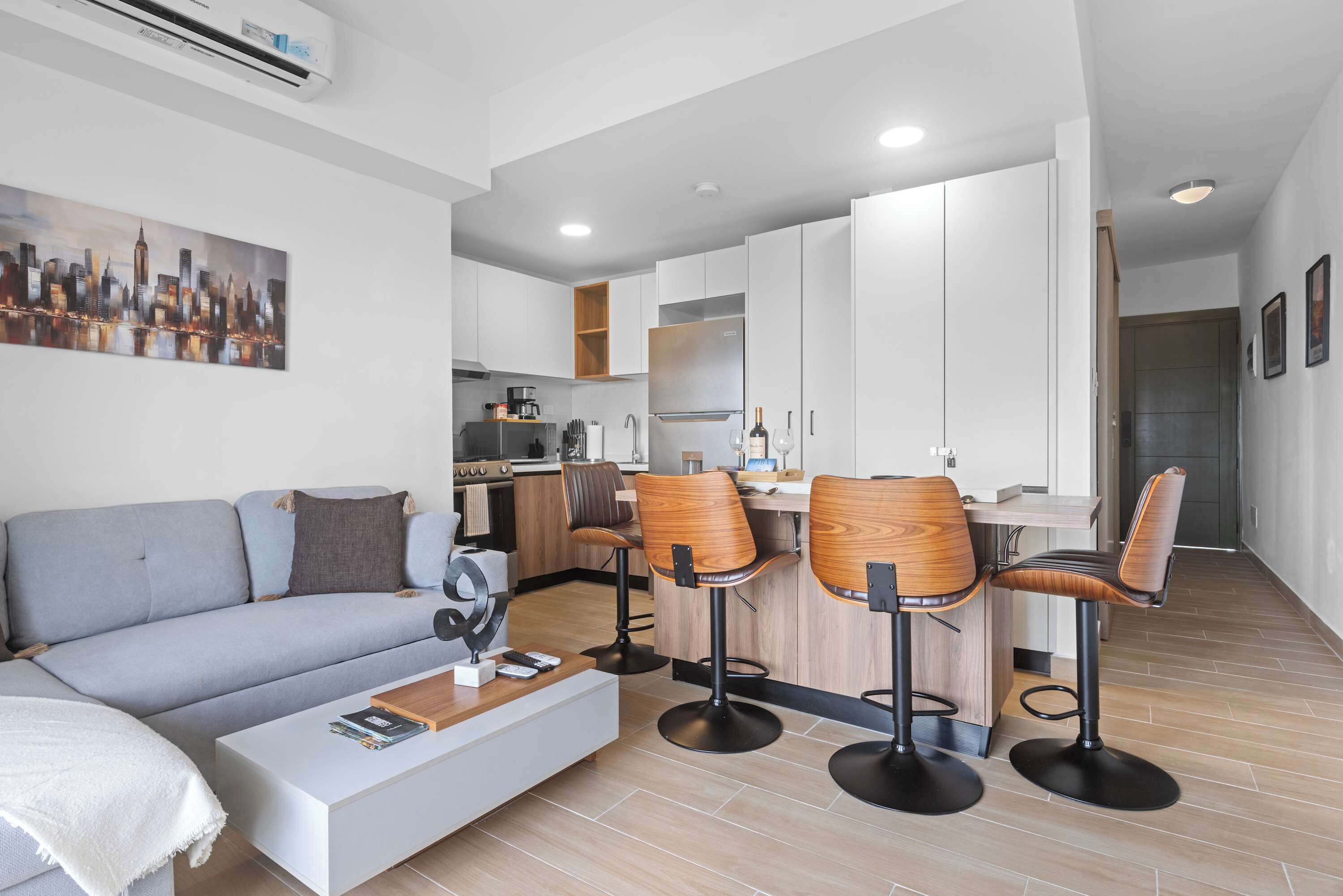A modern living room with a sofa, coffee table, and bar stools. A kitchen with white cabinets and a dining area is visible in the background.