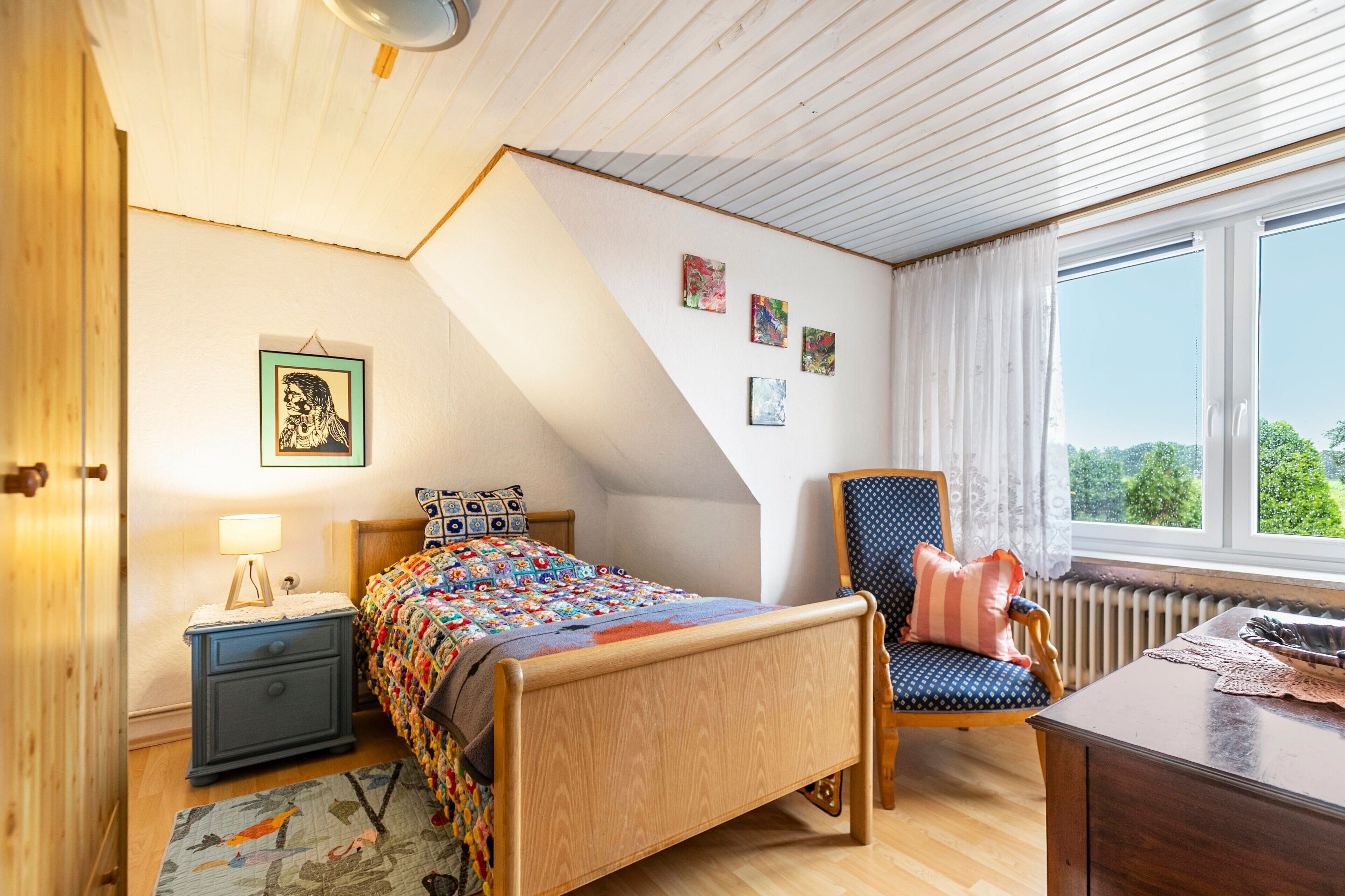 Ein Schlafzimmer mit einem hölzernen Bett, einem Nachttisch, einem Stuhl und einem Fenster mit Blick auf Grünflächen.