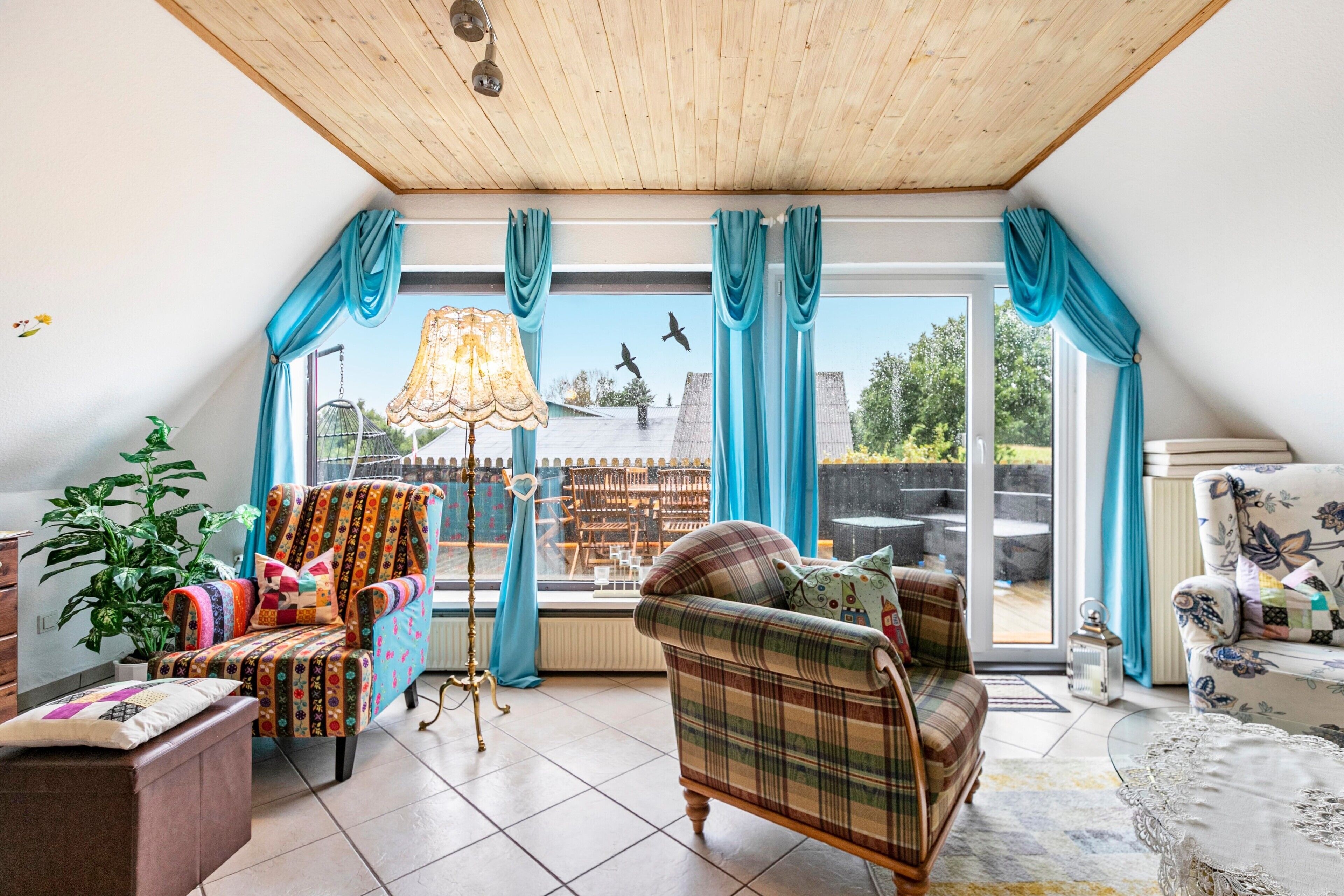 Une pièce chaleureuse avec un plafond en bois, une grande fenêtre avec des rideaux, un canapé, un fauteuil, une table et une lampe.