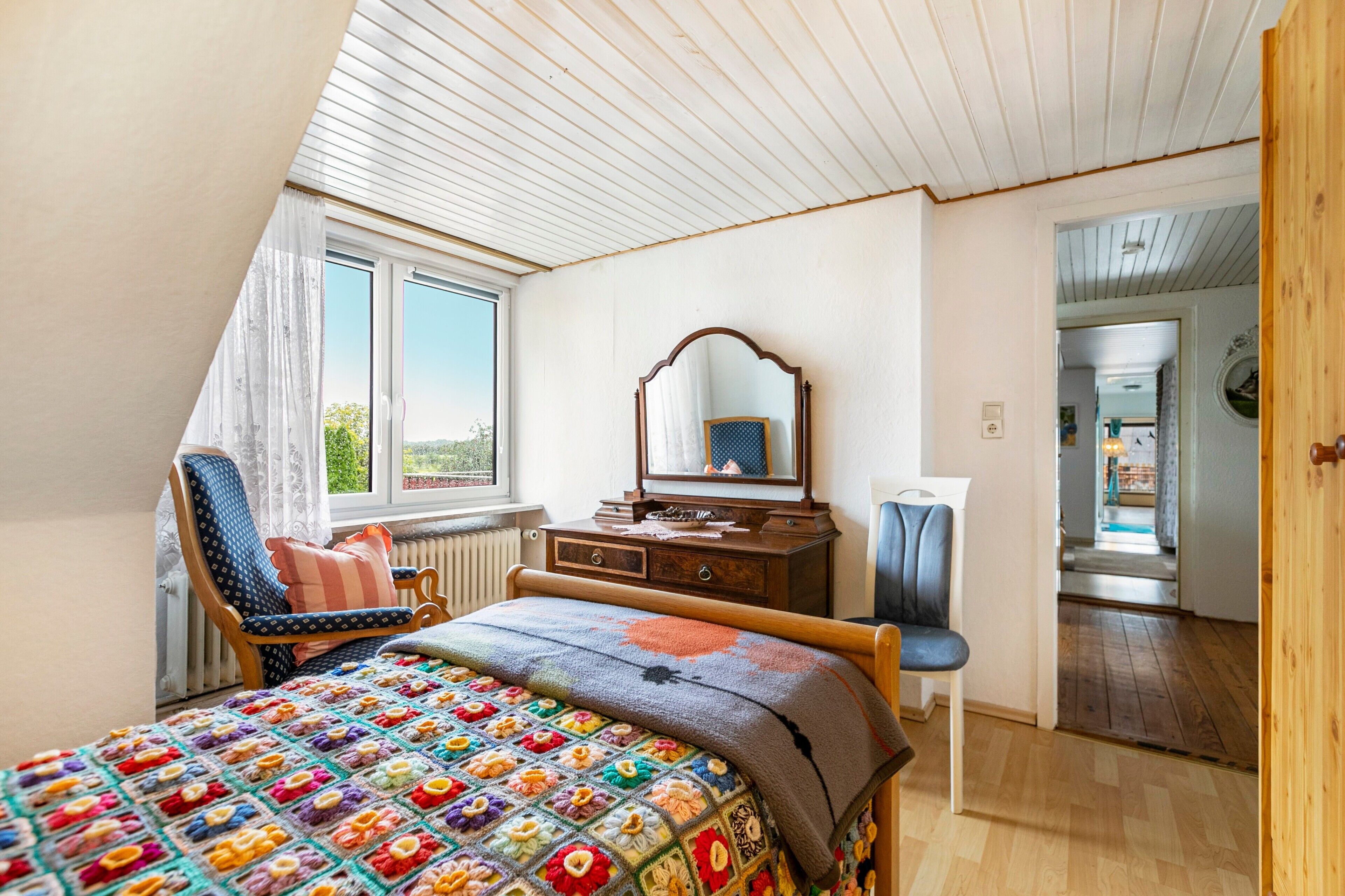 Quarto com teto inclinado, uma janela grande, cama com manta colorida, cômoda de madeira e uma cadeira.