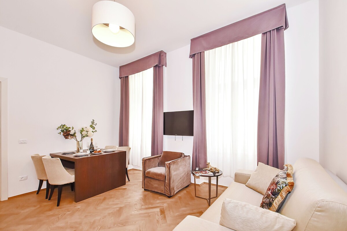 A modern living room with a dining area, large windows with purple curtains, a flat-screen TV mounted on the wall, and a wooden floor.