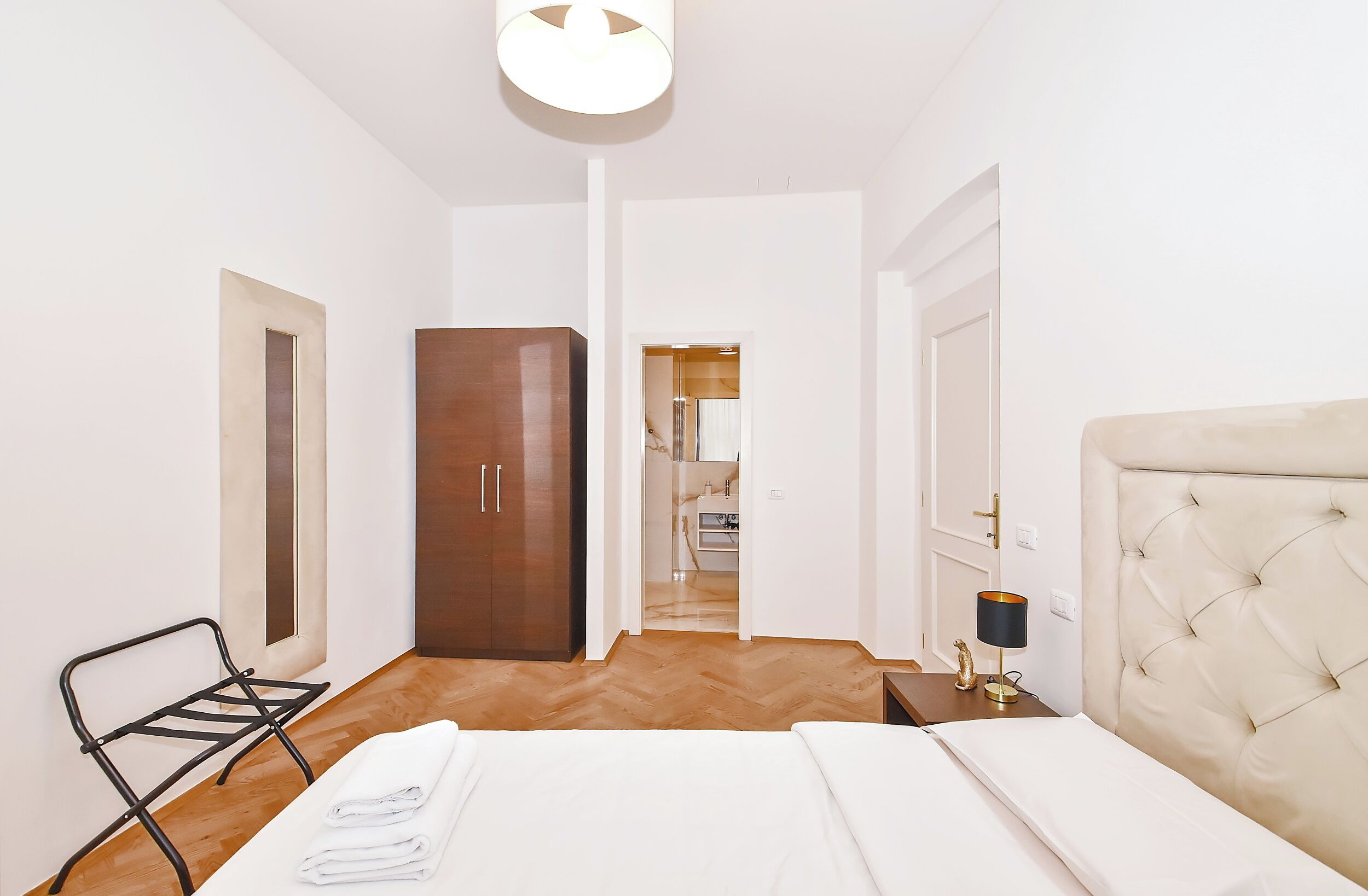 A hotel room with a bed, a wardrobe, a chair, a lamp, and a view into the kitchen.