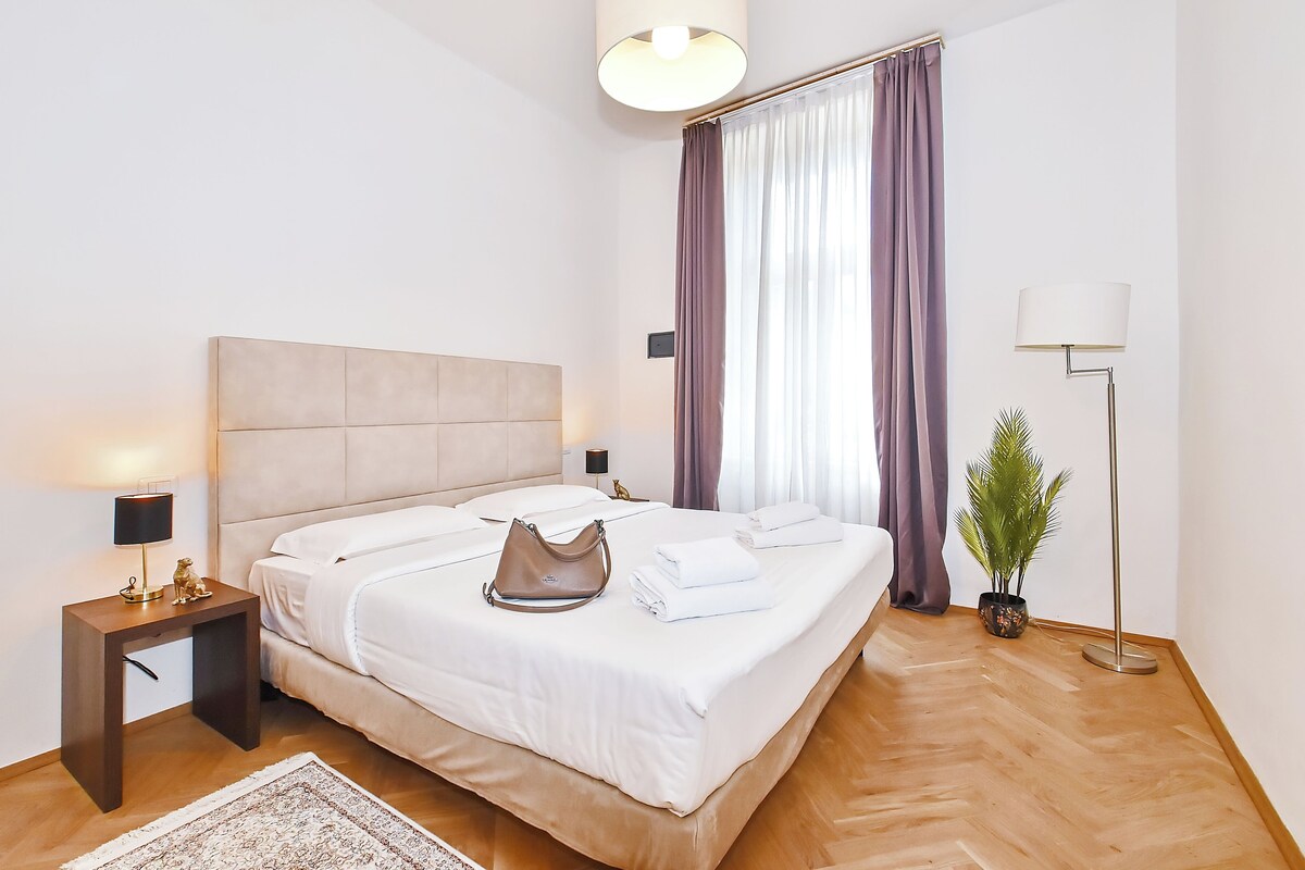 A hotel room with a bed, bedside lamps, a nightstand, a potted plant, and a floor lamp.
