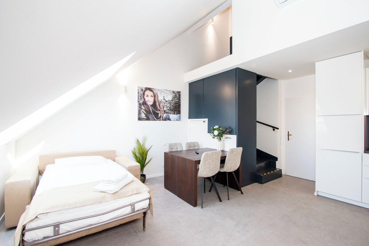 A modern bedroom with a bed, a desk, and a chair. There is a staircase leading to an upper level.