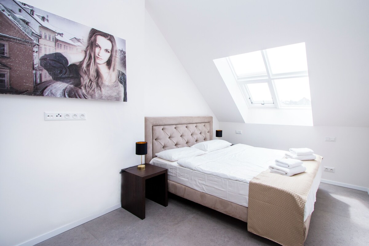 A bedroom with a large bed, a nightstand, a wall-mounted lamp, and a painting.
