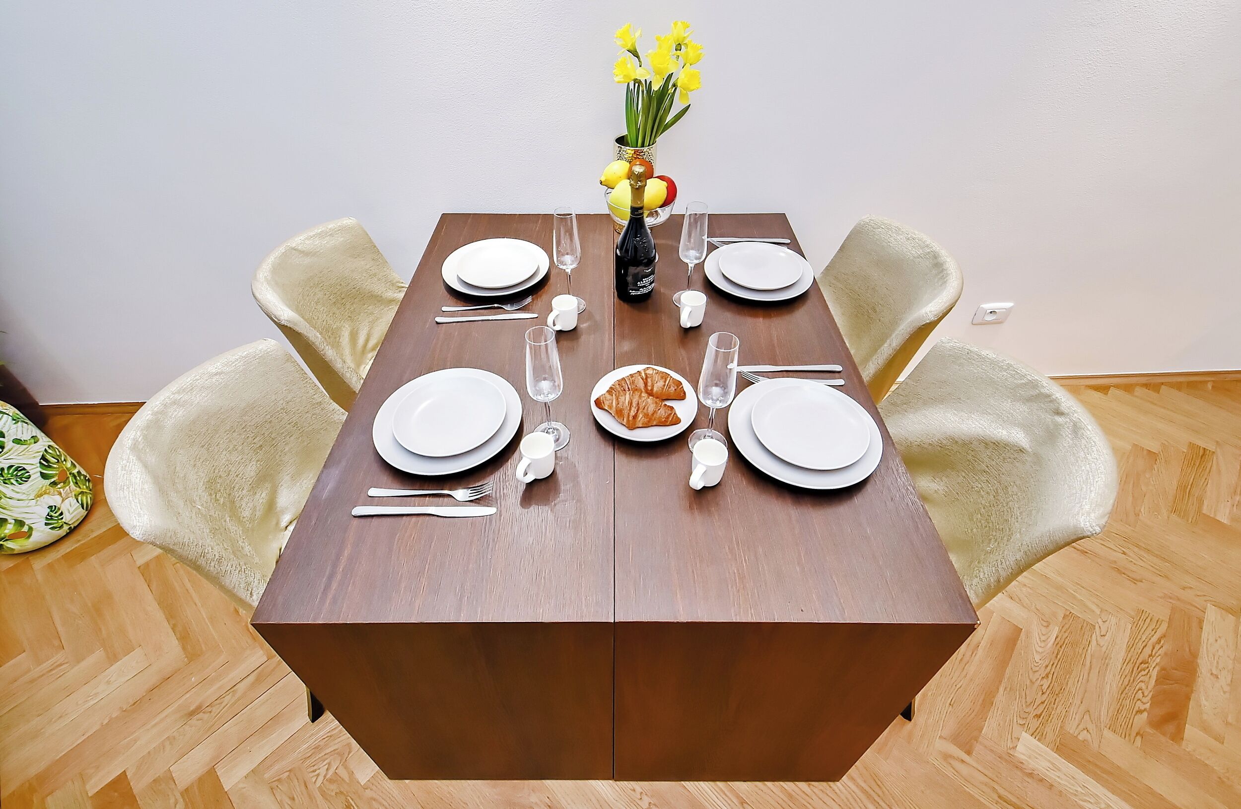 Ett dukat matbord med tallrikar, bestick, glas, en flaska, en vas med blommor och en croissant.