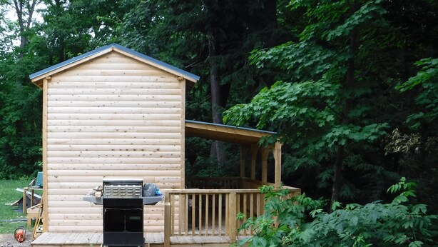 A wooden cabin with a deck, surrounded by dense forest. - Rustic Bunkie with Lake Front View! (Echo Bay)