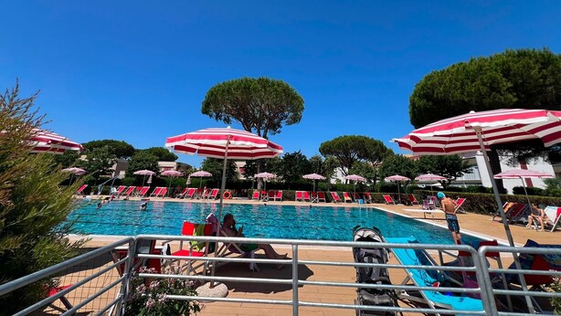 Una piscina amb ombrelles blaves i vermelles a ratlles, envoltada de gandules i arbres.
