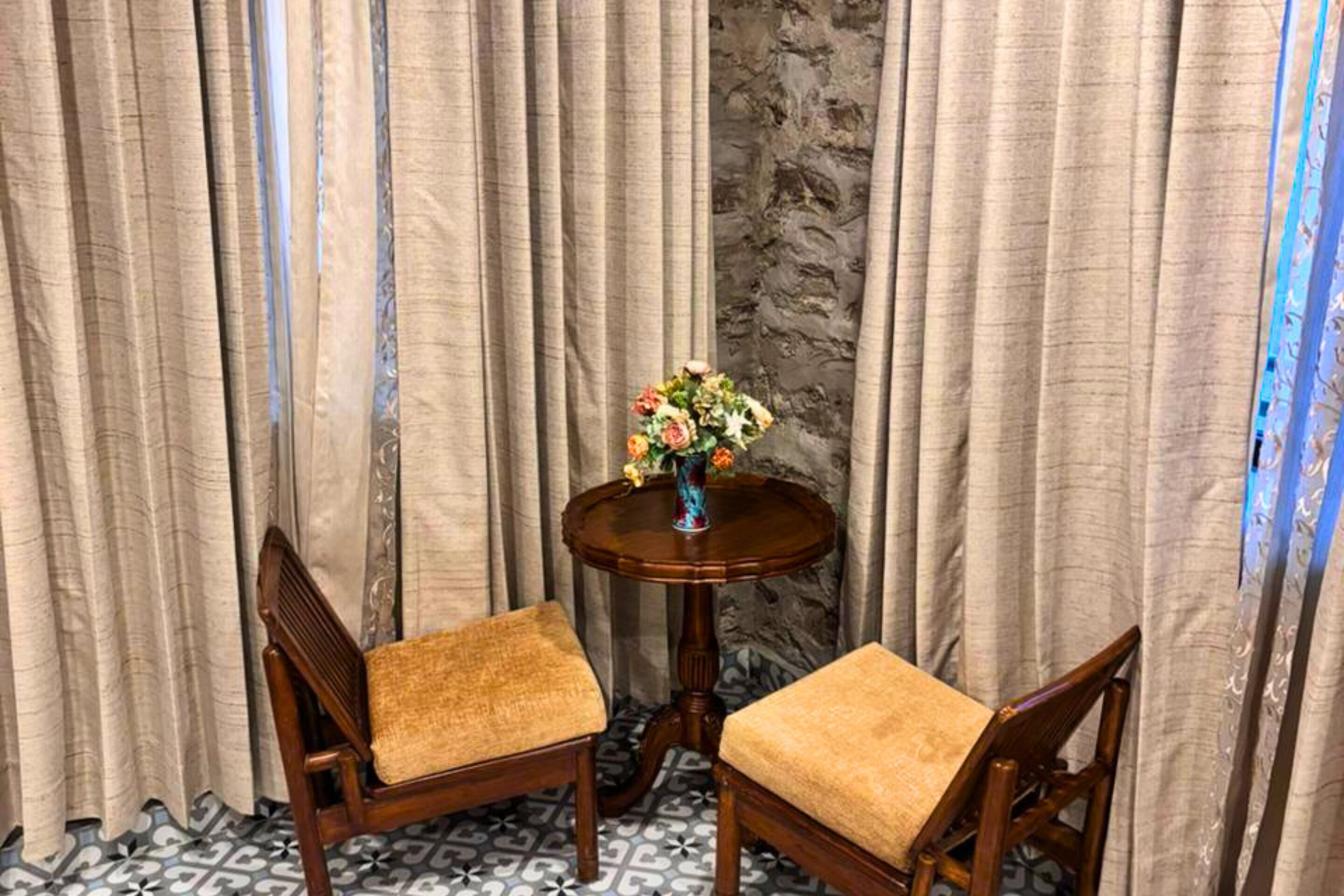 Ein Zimmer mit zwei Holzstühlen, einem runden Holztisch mit einer Blumenvase und Vorhängen.
