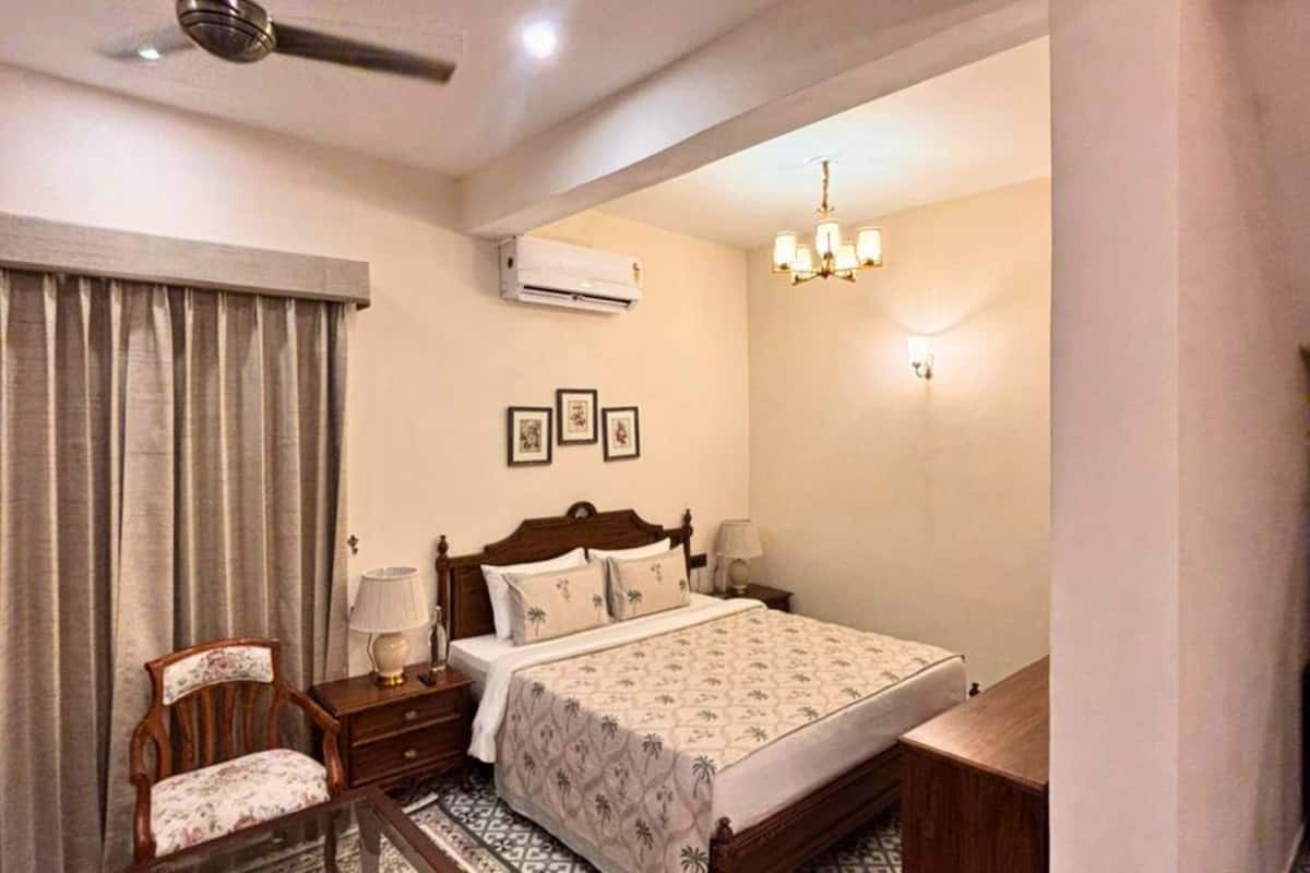 A bedroom with a bed, a chair, a nightstand, an air conditioning unit, and a chandelier.